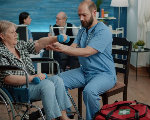 Medical assistant helping disabled woman with physical exercises in nursing home. Nurse giving assistance to senior patient in wheelchair while doing activity with dumbbells for recovery
